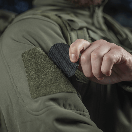 Person fäster M-Tac Patch Operator 3D PVC med Velcro på jacka, taktisk design, morale patch, 6x7 cm, av PVC.