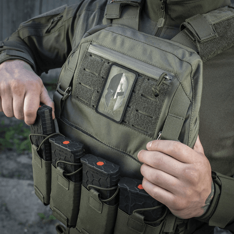 Man bär M-Tac Spartan Hjelm Patch, Cordura 500D, på militärväst, visar mod och styrka med Velcro‑fäste, vintagelook.