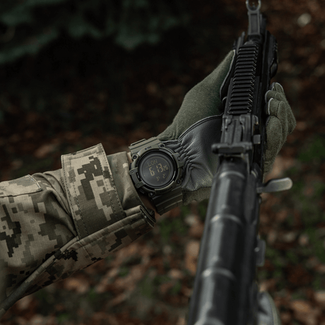 M-Tac Tactical Klocka med Kompass 2233 bärs av person i kamouflage med gevär, visar hållbarhet och funktionalitet i vildmarken.
