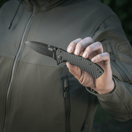 Person håller M-Tac Outdoor & Militär fällkniv - Type 7 med svart blad och ergonomiskt grepp, lämplig för taktisk användning.