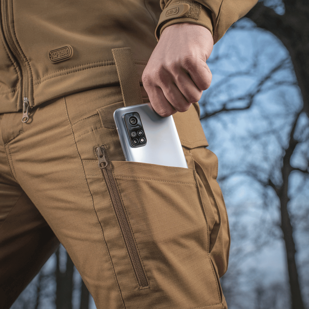 Mand iført M-tac Taktiske Bukser Conquistador Gen. I Flex med telefon i lommen, ideel til udendørsaktiviteter, lavet af Rip-stop Flex stof.