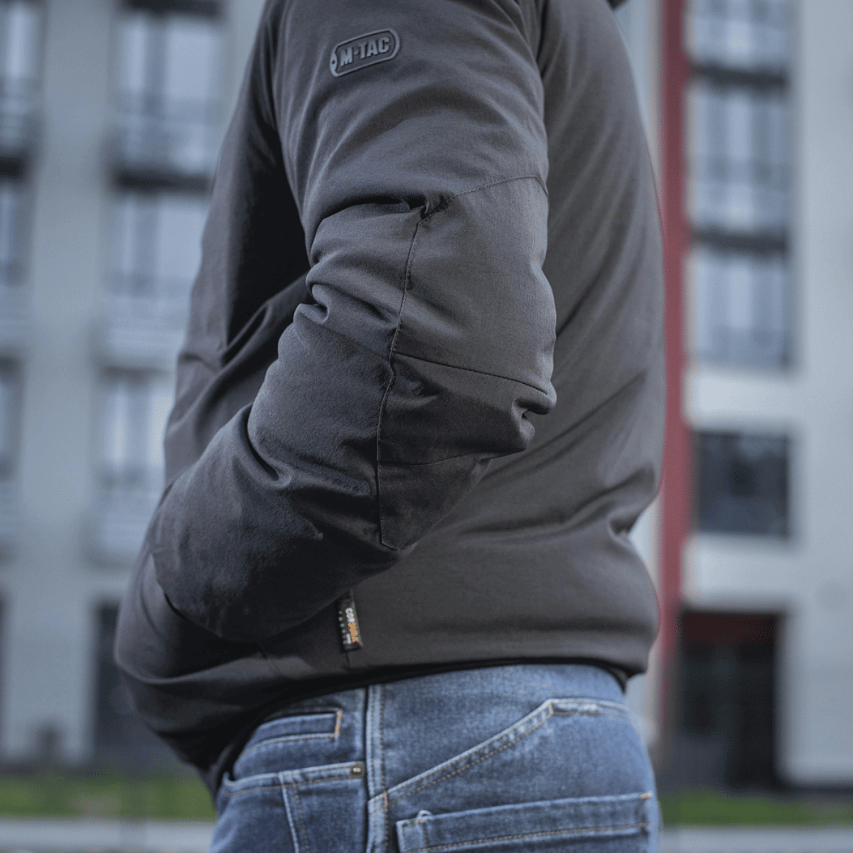 Person iført M-Tac Rubicon Jakke Gen. II – Slidstærk og varm bomberjakke, med bygninger i baggrunden, set fra siden.