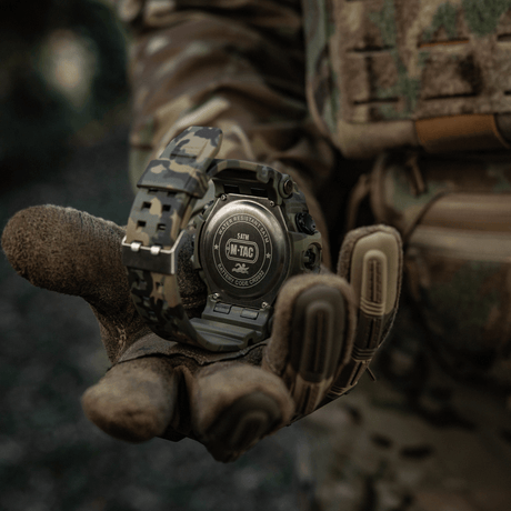 M-Tac Tactical Klocka 2037 i camouflagedesign, tålig upp till 5ATM, hålls av en taktisk handske i skogsmiljö.