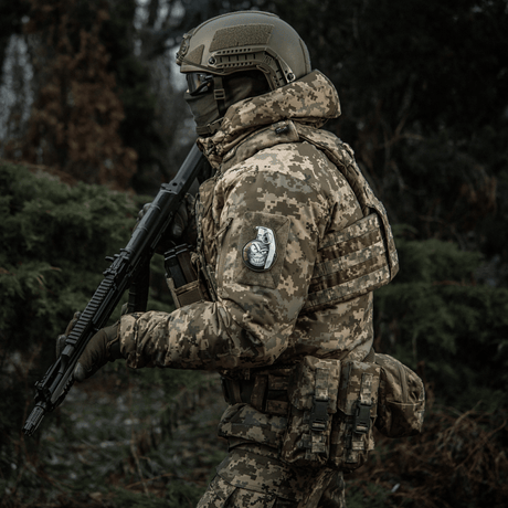 Soldat i kamouflageuniform med M-Tac Patch M67 granat 3D PVC på ärmen, beväpnad i skogen.