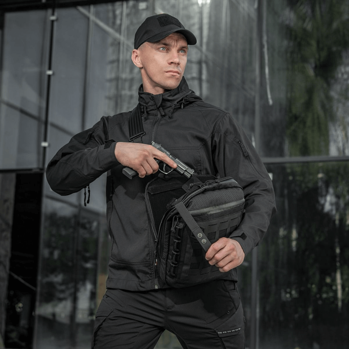 Person holder M-Tac Tablet Bag Laser Cut Hex og afslører skjult våben foran glasfacade, iført sort jakke og hue