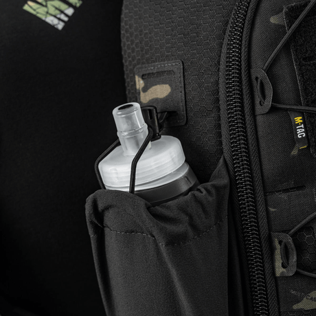 Sort rygsæk med netlomme og hvidt, rund låg til drikkeflaske, perfekt til udendørs aktiviteter. Camouflagedetaljer og lynlås synlige.