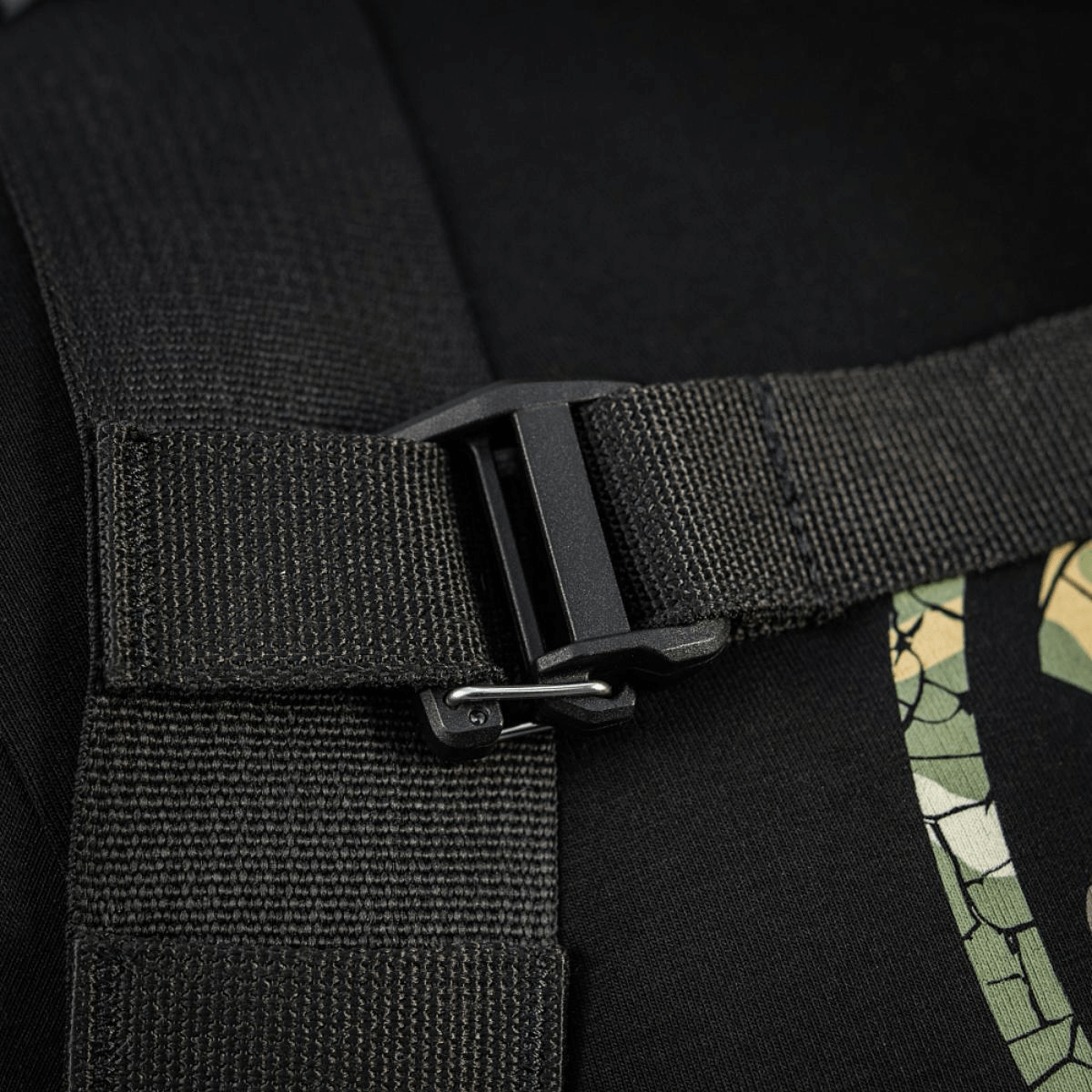 Nærbillede af sort stropspænde på rygsæk med camouflage-trykdetalje, robust konstruktion og sikker lukning.