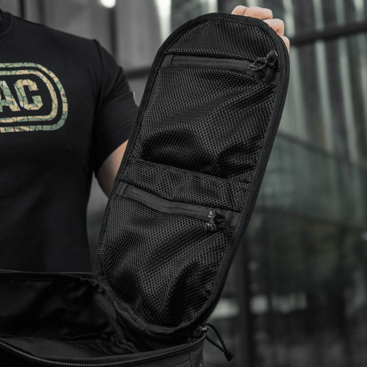 Sort taske med netlommer og lynlåse åbnet på en moderne bybaggrund, betonet af en person i sort trøje.