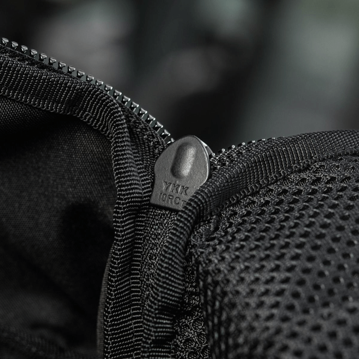 Nærbillede af sort lynlås på rygsæk med netdetaljer, der viser YKK-mærket, fokus på holdbarhed og kvalitet.