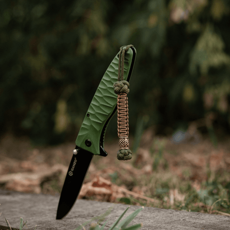 M-Tac Knife Lanyard Celtic: Flettet paracord lanyard på grøn kniv i naturlig udendørs omgivelser, stilfuldt og praktisk tilbehør.