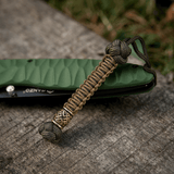 M-Tac Knife Lanyard Celtic i flettet paracord ved siden af en grøn lommekniv på træoverflade