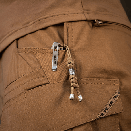 M-Tac Zeus Stainless Steel Lanyard i lommen på brune bukser med dekorativ perle og flettet paracord, nem adgang til kniv eller udstyr.