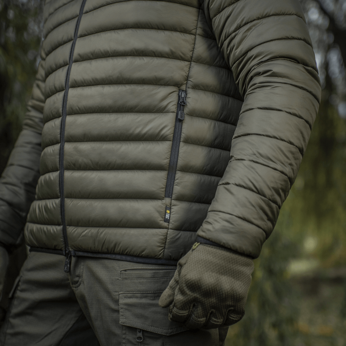 Mand i M-Tac Stalker Gen.III Jakke i skovmiljø, viser detaljeret syning og lynlås, perfekt til overgangssæsonen.