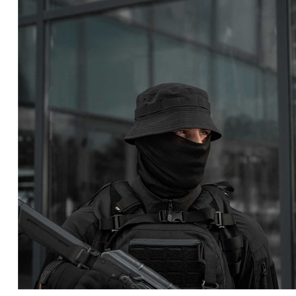 Mand iført taktisk tøj med sort hat, maske og rygsæk foran glasbygning.