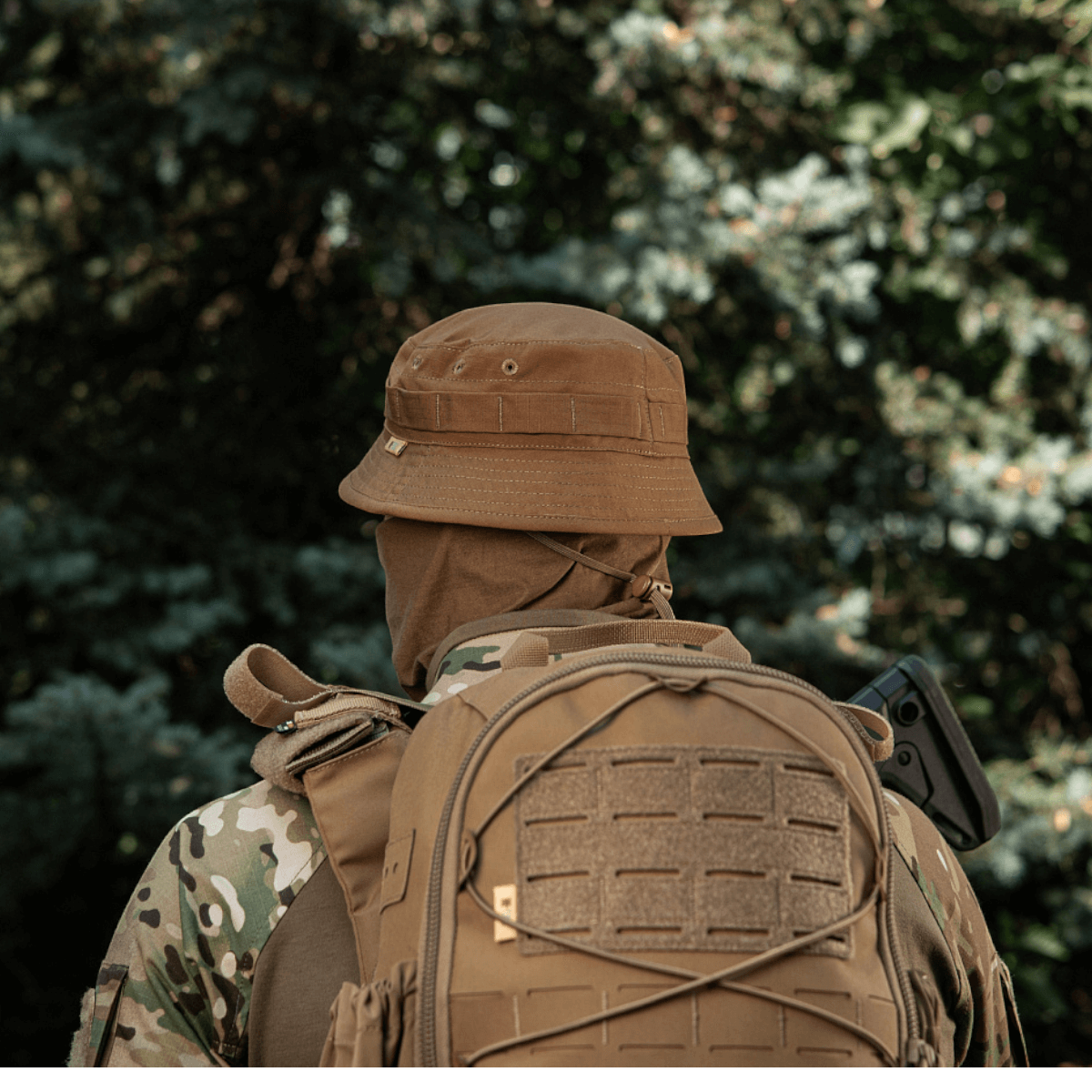 Person i taktisk camouflagedragt og rygsæk iført M-Tac Panamahat Gen. II Flex Rip-Stop i skovmiljø.