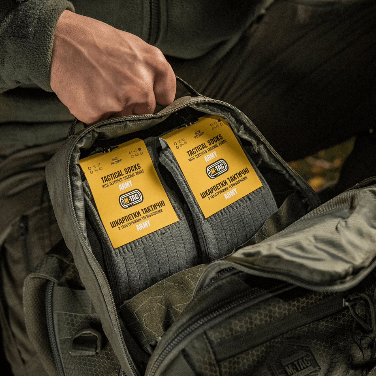 Person pakker M-Tac Sokker Tactical Army i en rygsæk til udendørs brug, viser produktemballagen med fokus på komfort og holdbarhed.