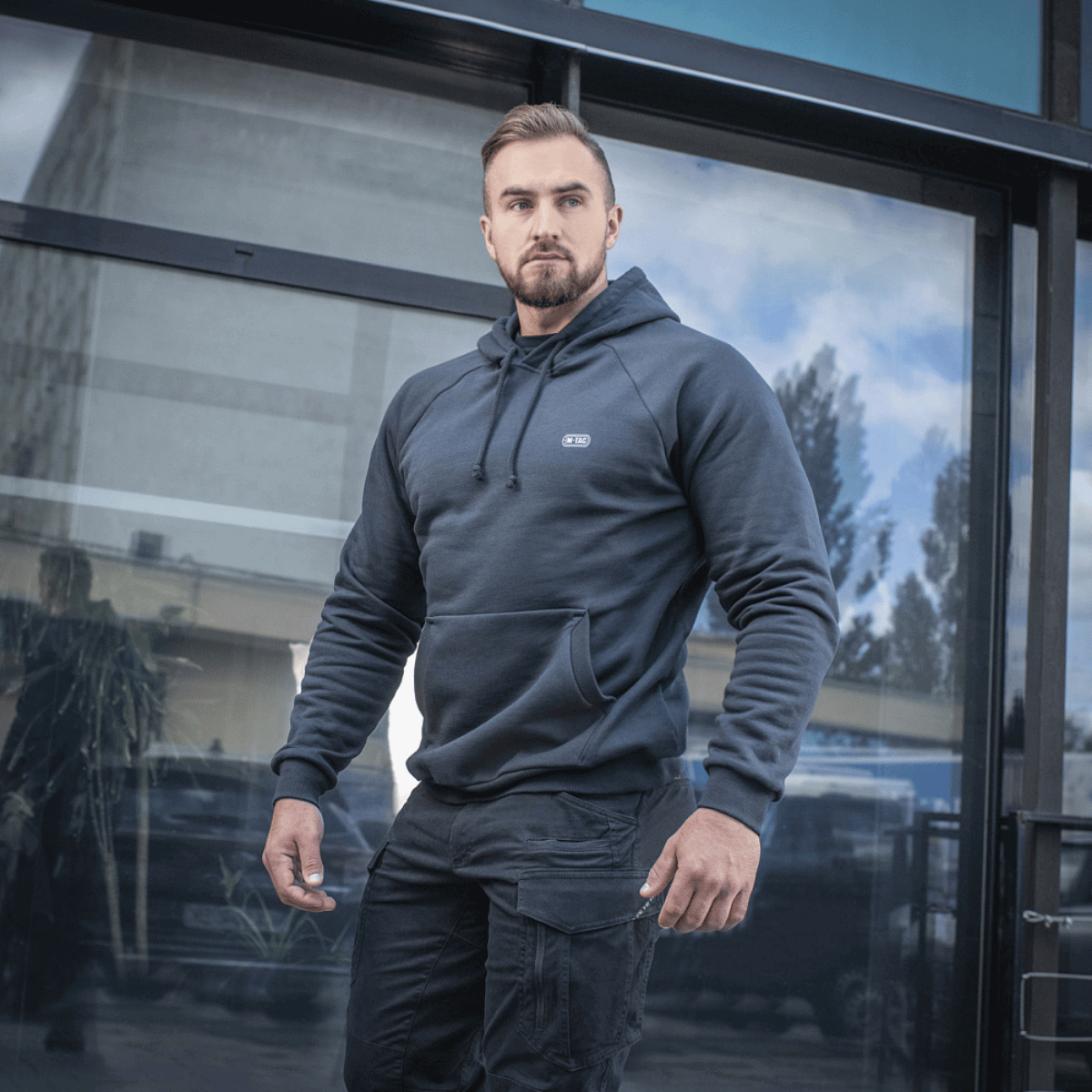 Mand iklædt M-Tac Hættetrøje Cotton Raglan foran glasbygning, bærer afslappet og komfortabelt look, perfekt til hverdagsbrug.