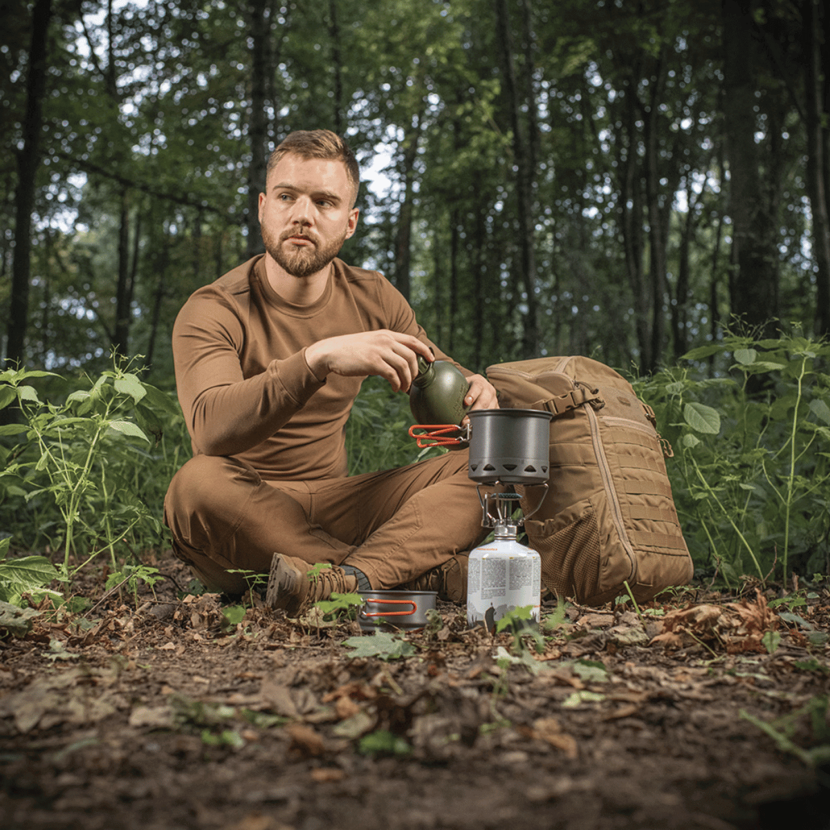 Mand i skov bruger M-Tac Individuelt Kogesæt til Brænder til camping, sidder ved komfur med rygsæk.