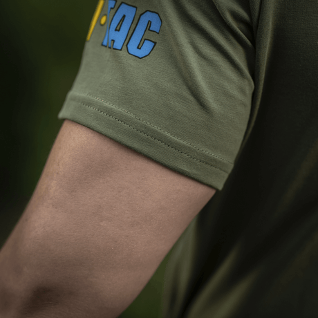 Close-up af ærmet på M-Tac T-Shirt Quadcopter med logo i blå og gul på olivengrøn baggrund, viser bomuld og elastan blanding.