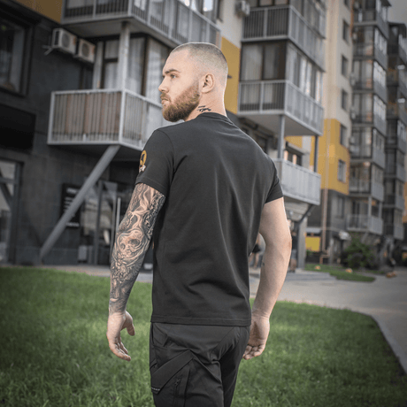 Mand bærer en M-Tac T-shirt Black Sea Expedition foran moderne bygninger