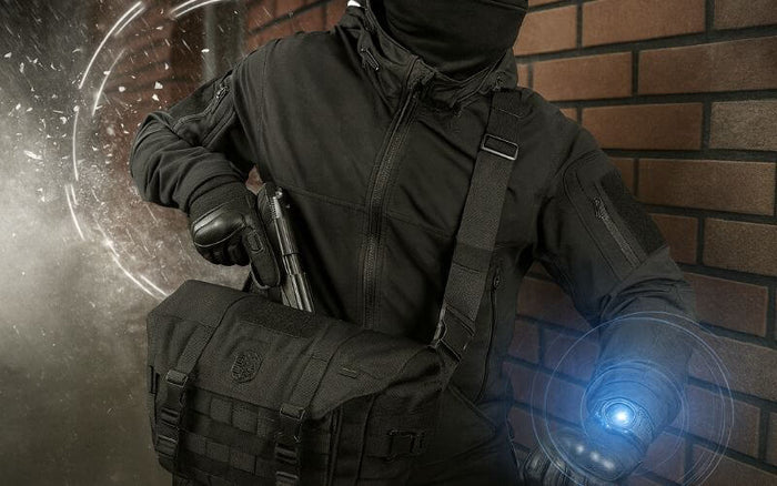 Taktisk klædt person med skuldertaske og lommelygte, står op ad murstensvæg, klar til mission. Effektivitet og beredskab.